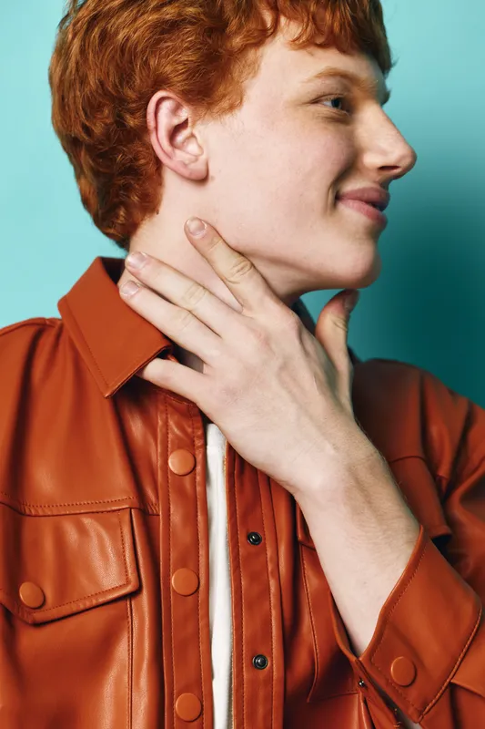 Young man with red hair wearing a brown leather jacket smiles looking to the side, hand touching neck against blue background in casual fashion portrait.
