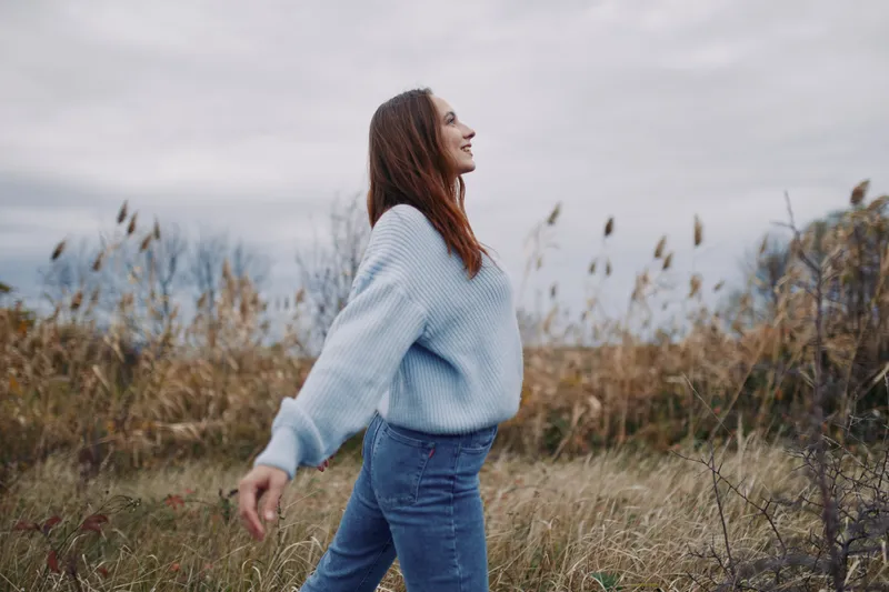 A candid portrait of a young woman walking in a windswept field, smiling with natural ease, evoking authenticity and quiet confidence in an unposed, believable lifestyle moment.