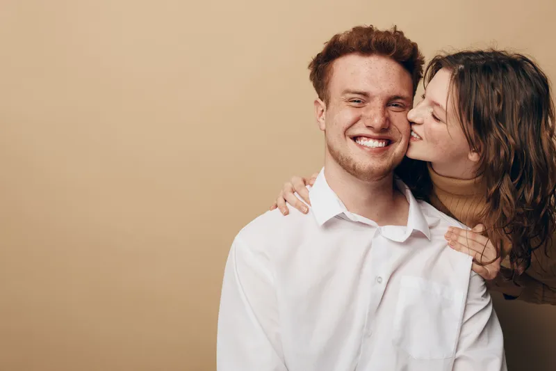 Happy young couple in casual clothes share affectionate moment, woman kissing man on cheek. Warm beige background, natural light, concept of love, relationship, and intimacy.