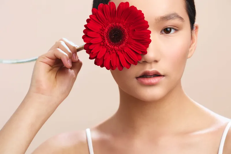 Asian woman is featured in a closeup portrait, holding a bold red flower near her face. The composition emphasizes elegance, calm, and refined beauty with soft studio lighting.