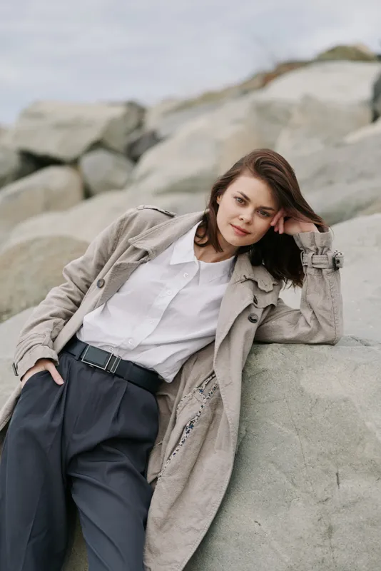 Young woman in casual outfit leans on large rocks outdoors, wearing coat and white shirt. Natural light highlights relaxed mood and neutral tones in a scenic rocky environment.