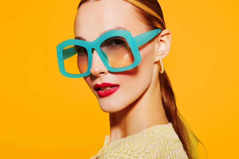 Stylish woman with red lipstick wearing oversized turquoise sunglasses and yellow sweater poses against bright yellow background. Concept of fashion, summer style, and confident attitude.