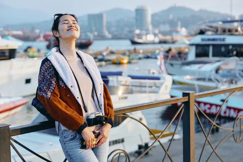 Woman by a waterfront harbor in casual denim jacket and scarf, enjoying sunny outdoor scene among boats, rails and distant cityscape, relaxed mood and fashion style