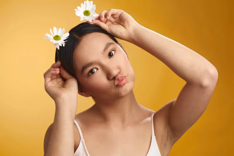 asian woman with daisies in her hair stands before a warm backdrop, creating a bright portrait that radiates cheerfulness, natural charm, and a friendly, inviting mood.