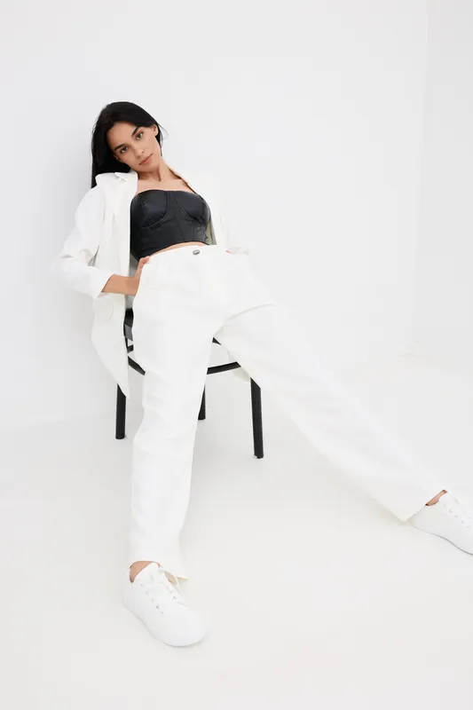 Stylish woman wearing white pants and blazer with black top, sitting relaxed on chair against plain white background, casual modern fashion portrait.