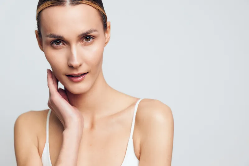 Young woman with clear skin gently touches her cheek while looking at the camera, posing in a tank top against a pale gray studio backdrop, with soft studio lighting and calm mood.