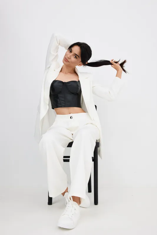 Young woman sitting on chair pulling her long dark hair, wearing white suit and black corset top, studio background with modern casual fashion style.