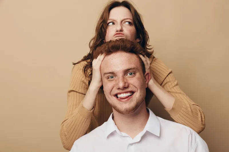 Happy redhead man smiles while woman in beige sweater playfully holds his head from behind. Studio portrait on beige background. Concept of friendship, fun, and playful relationship.