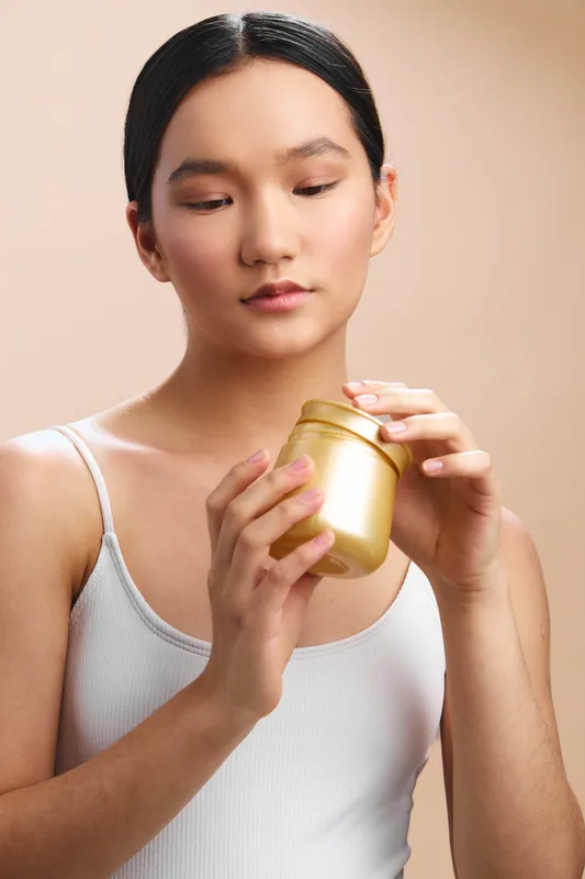 asian woman calmly examines a gold cosmetic jar in a minimalist studio, presenting a fresh skincare moment that conveys elegance, focus, and everyday beauty in a serene setting.