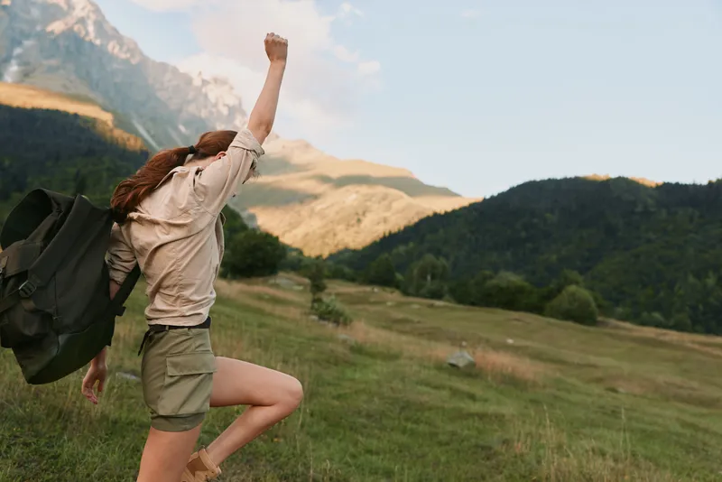 A woman in casual clothes hikes up a grassy mountain trail with a backpack, raising her arm in triumph. The scenic landscape features green hills and a clear sky.