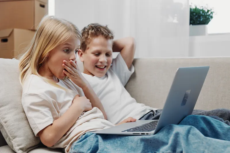 Two children sit on a couch watching a laptop screen, girl surprised and boy laughing, casual home setting with natural light. Concept of childhood, technology, entertainment and family.