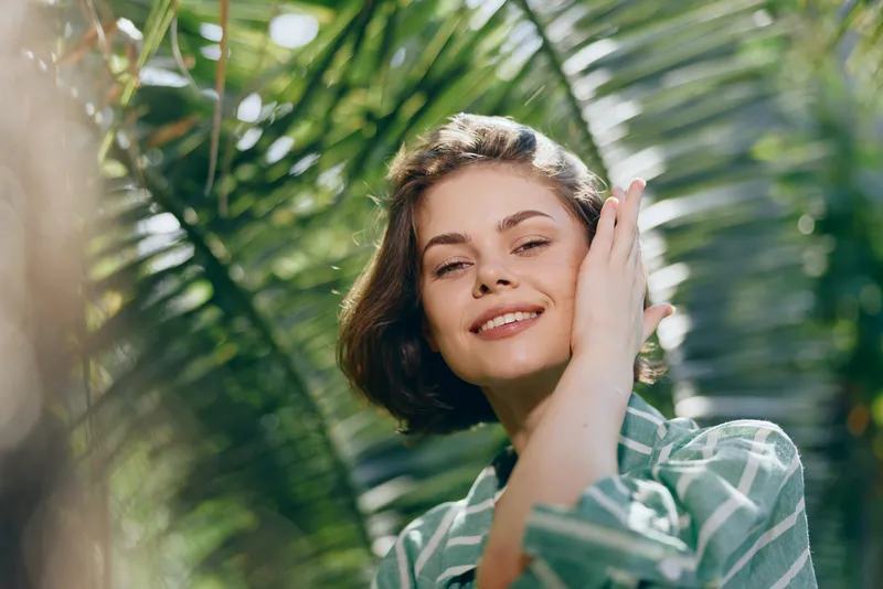 A cheerful woman relaxes outdoors amid lush greenery, smiling warmly as sunlight highlights her face.