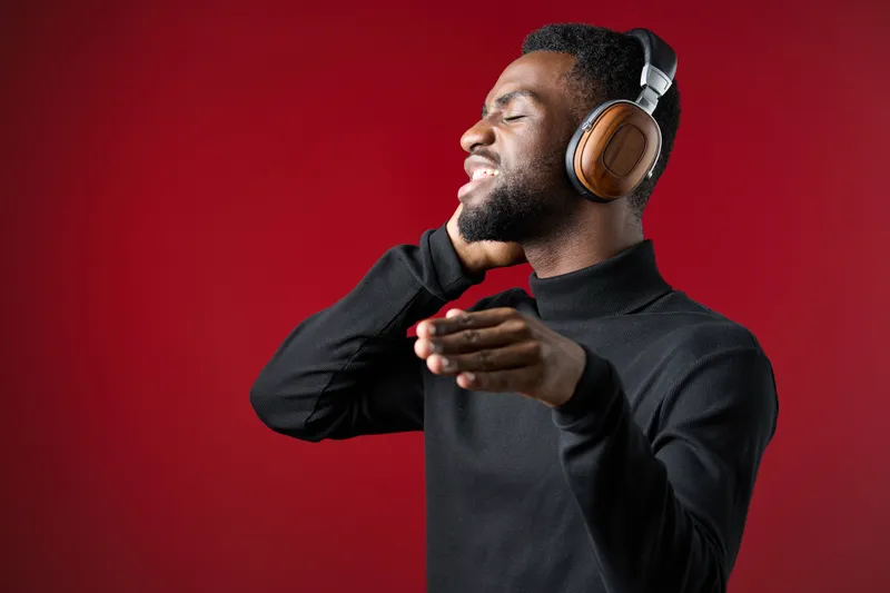Young African man enjoying music with headphones against a vibrant red background, expressing joy and emotion through his gestures