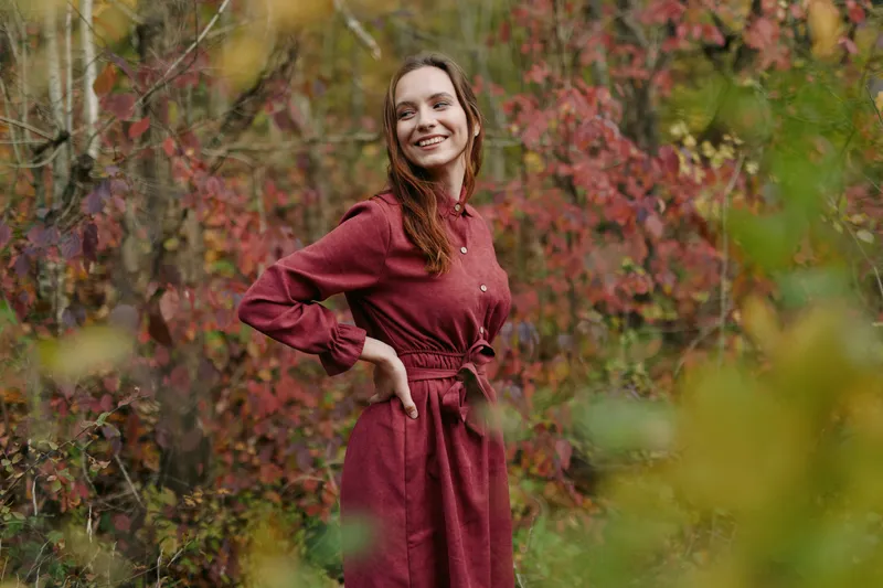 Genuine portrait of a smiling woman in a simple red dress among autumn foliage, capturing authentic natural expression and an honest connection with the serene outdoor environment and season.