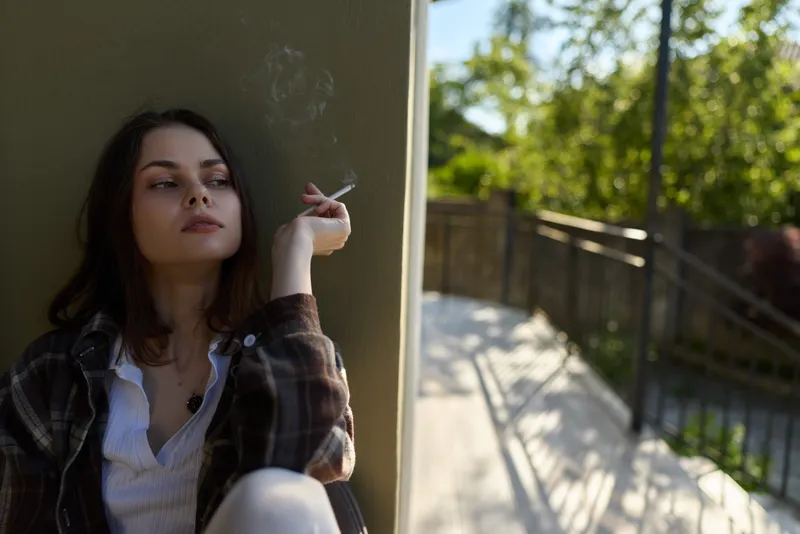 A pensive woman leans against a concrete wall, smoking outdoors in casual clothing. A quiet, moody moment capturing lifestyle, solitude, and introspection