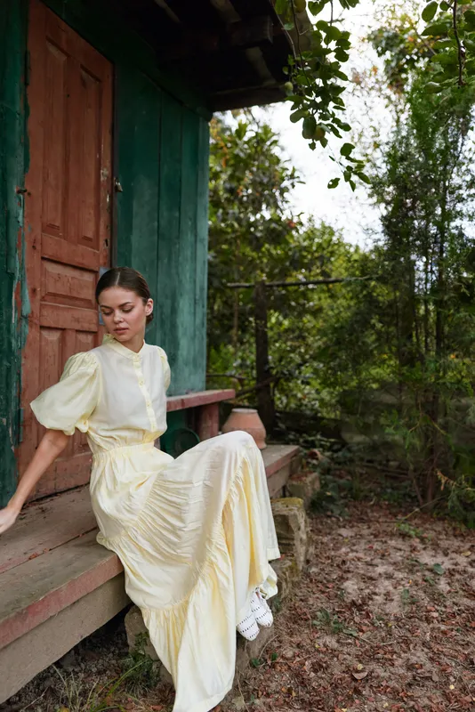 A woman in a pale yellow dress sits on wooden steps outdoors, surrounded by greenery, creating a calm rustic portrait bathed in natural light.