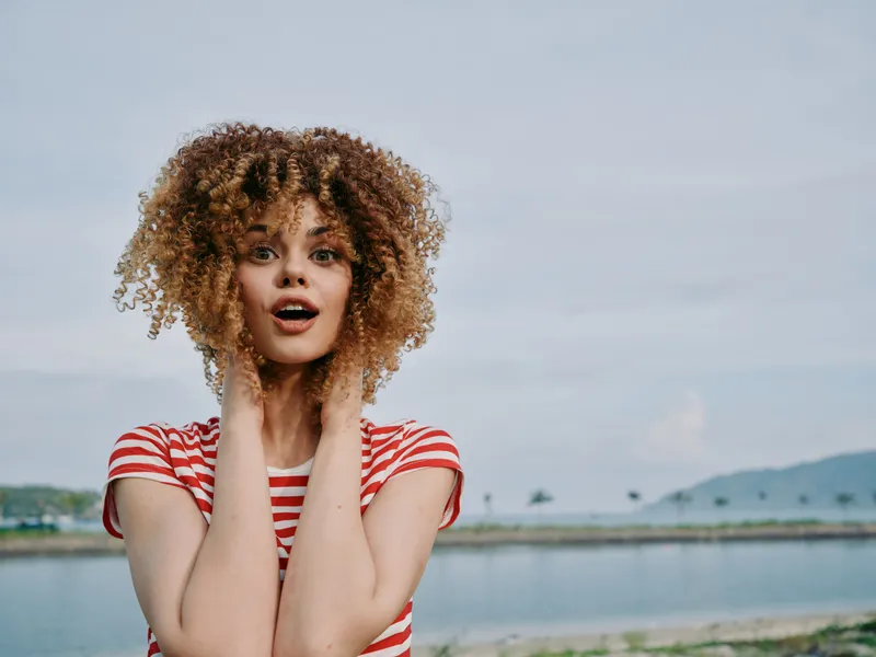 Portrait of a young woman with voluminous curly hair wearing a red and white striped shirt, outdoors beside a calm lake, her eyes wide and mouth open in a surprised expression.