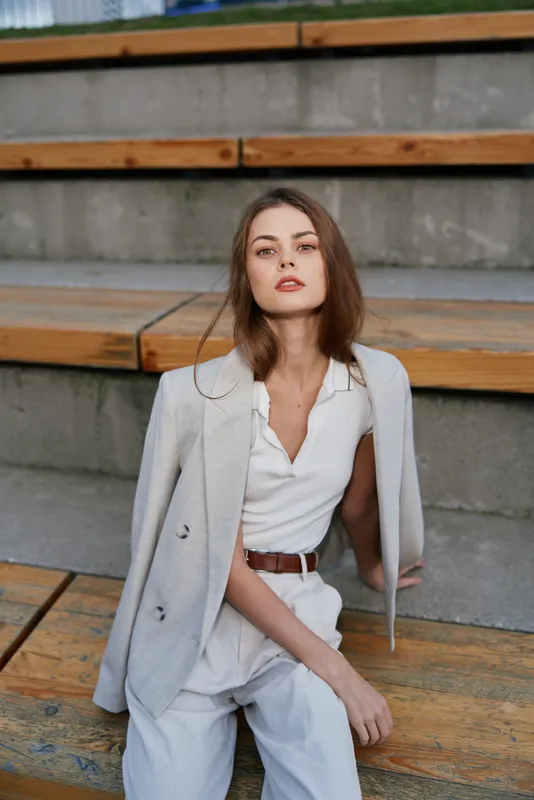 confident woman in white outfit and light jacket seated on wooden urban stairs with relaxed expression business fashion concept