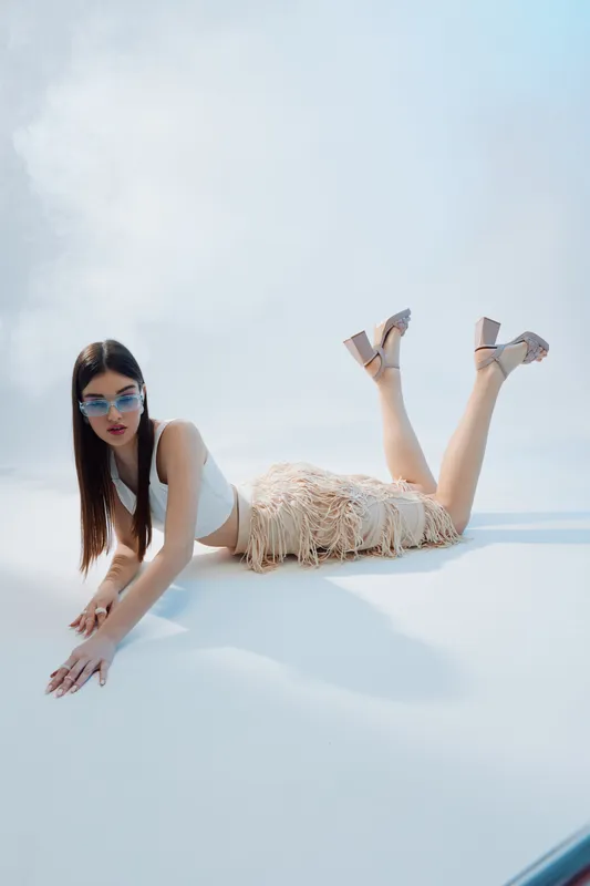 Fashionable woman in stylish sunglasses and fringed skirt lies on white surface under bright sky. Concept of beauty, fashion, summer, and modern lifestyle with minimalistic background.