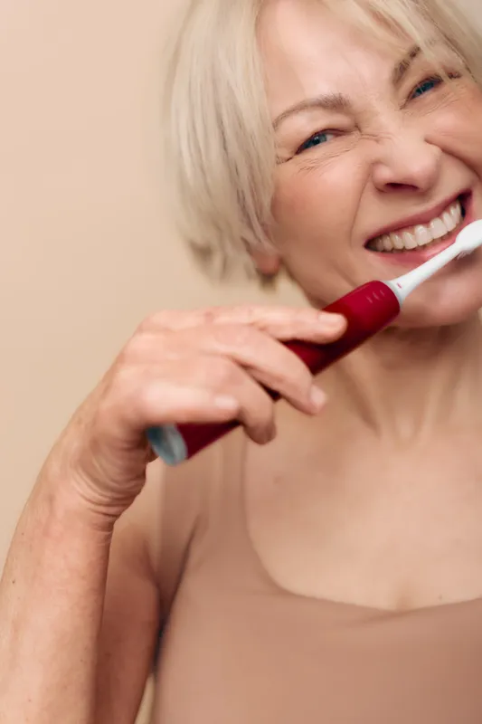 Smiling mature woman with short blonde hair brushing teeth using electric toothbrush. Close-up on beige background. Concept of dental care, hygiene, and healthy lifestyle for seniors.