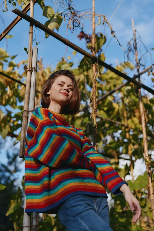 Woman in colorful striped sweater smiling outdoors in a garden portrait, casual young female standing by trellis with vines and blue sky, autumn light and relaxed pose.