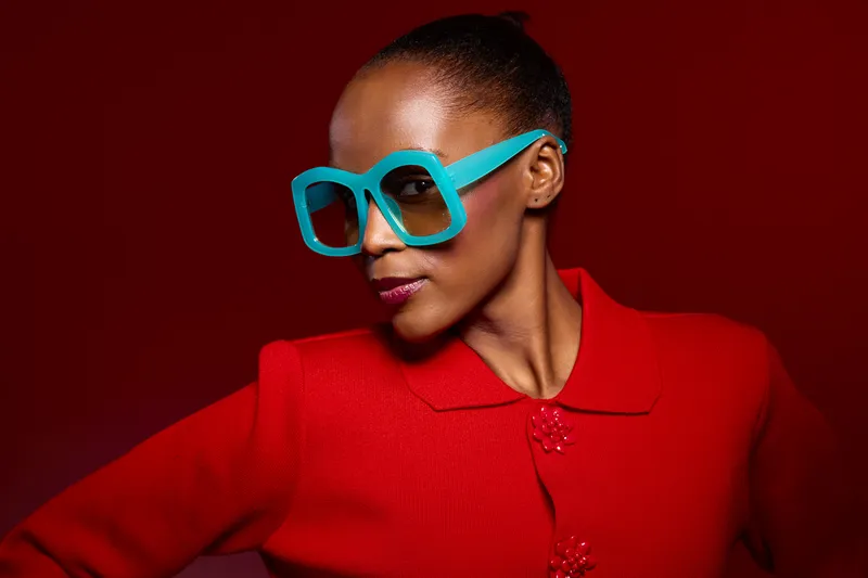 Stylish dark skinned African woman wearing bright blue oversized glasses and red textured jacket poses confidently against dark red background. Concept of fashion, portrait, and bold accessories.