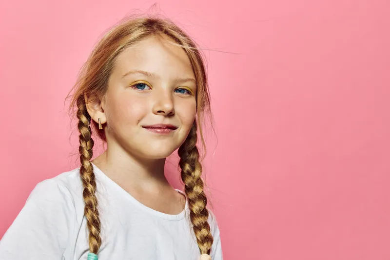 Smiling young girl with blonde braided hair wearing a white shirt against a pink background. Studio portrait with soft lighting and positive mood.