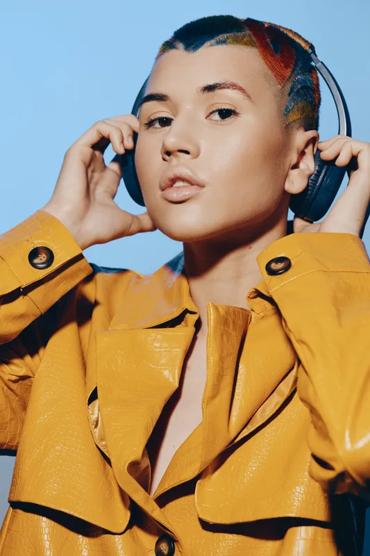 Woman with headphones wearing yellow leather coat adjusting music device, stylish short hair, bright blue background, modern urban fashion, confident expression.