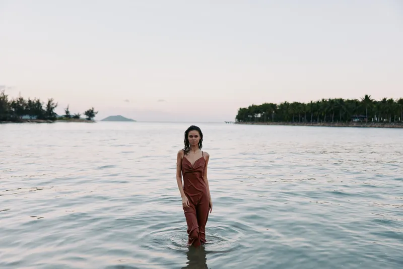 A lone woman stands waist deep in calm sea water at dusk, wearing a rust red dress. Soft light frames the distant horizon, creating a serene, cinematic coastal portrait.