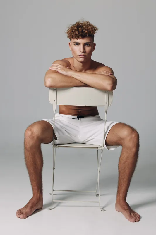 Young man with curly hair sitting barefoot on chair backwards wearing white shorts against gray background. Casual pose with arms resting on chair back and serious facial expression.