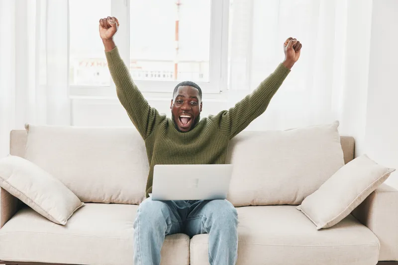 Happy man celebrating success at home, wearing a green sweater, sitting on a sofa with a laptop, conveying excitement and joy in a bright interior