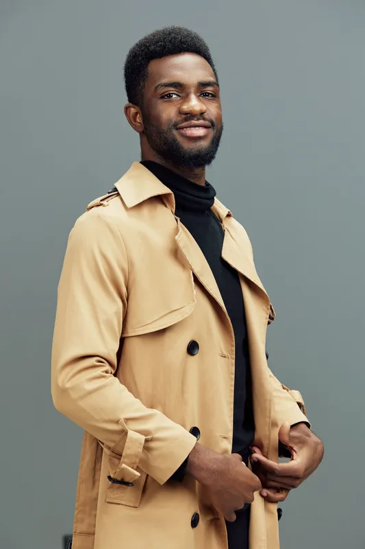 Stylish young African man wearing a beige trench coat and black turtleneck, smiling confidently against a gray background