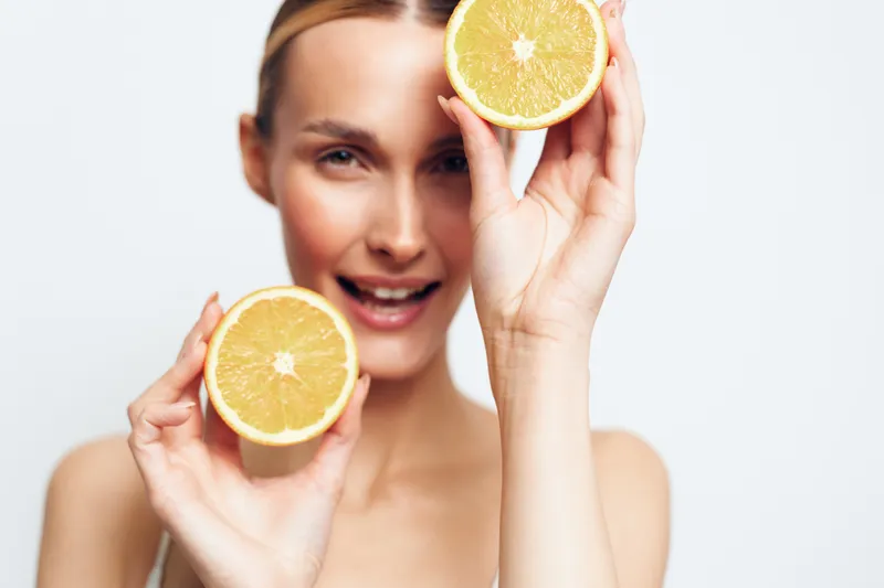 Young woman with clear skin holding two halves of fresh orange in hands, smiling on white background. Concept of healthy lifestyle, natural beauty, and vitamin C benefits.