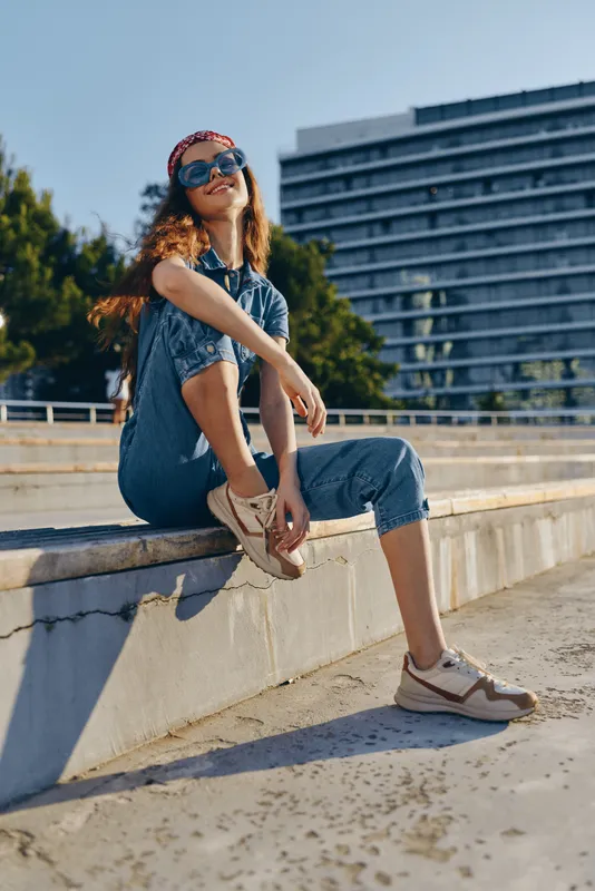 Lifestyle woman in modern boho-western denim street style sits confidently outdoors wearing sunglasses and sneakers with film color tones enhancing a casual urban vibe.