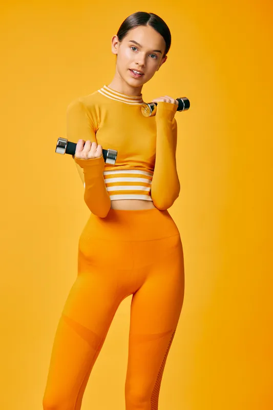 Fitness woman in a bright orange athletic set holds dumbbells against a vivid orange backdrop, performing bicep curls with focus and energy, showcasing strength, balance and a dynamic workout
