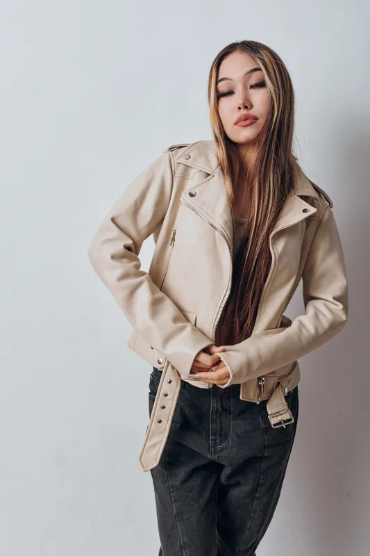 Young woman with long braided hair wearing beige jacket and black jeans standing against light background. Casual fashion style with confident pose and neutral expression.
