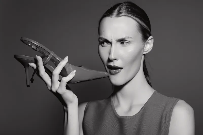 A pale white woman with slicked hair holds a high heel shoe near her face, expressing confusion and surprise. Studio portrait in black and white, concept of fashion and emotion.