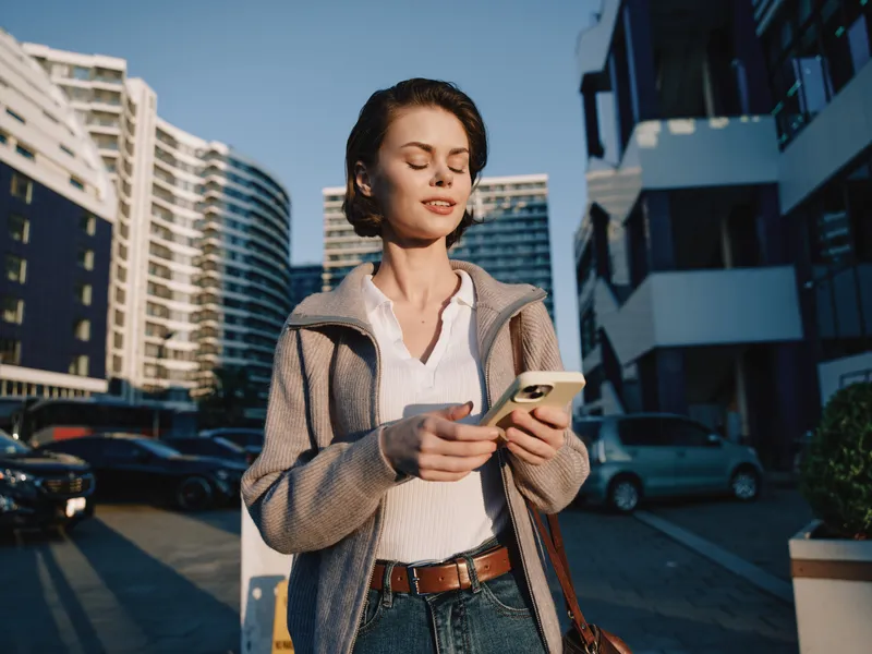 A woman is seen texting in an urban outdoor setting. She smiles softly and appears engaged, creating a positive and dynamic atmosphere in a modern street scene.