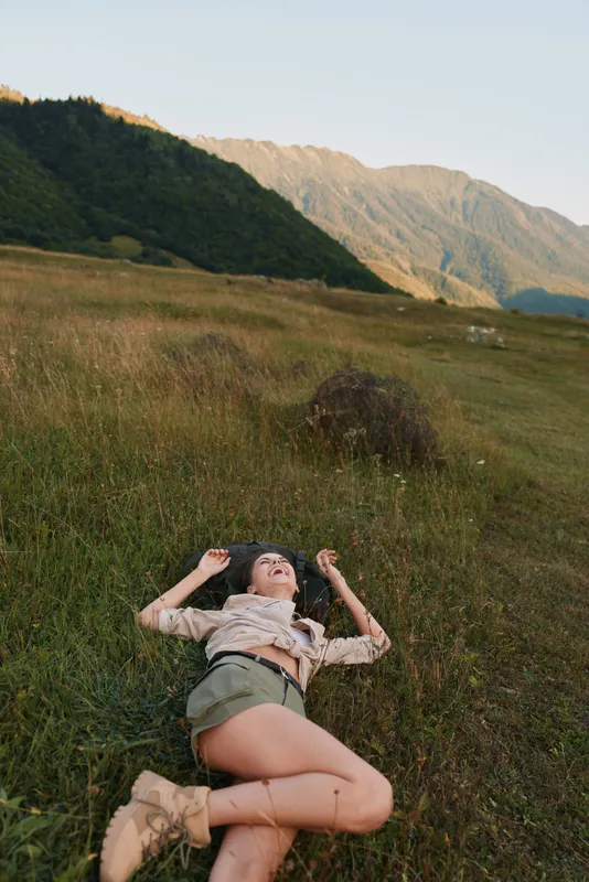 Young woman with long hair lies on grassy hill in casual outfit enjoying nature and relaxation during golden hour in a mountainous landscape.