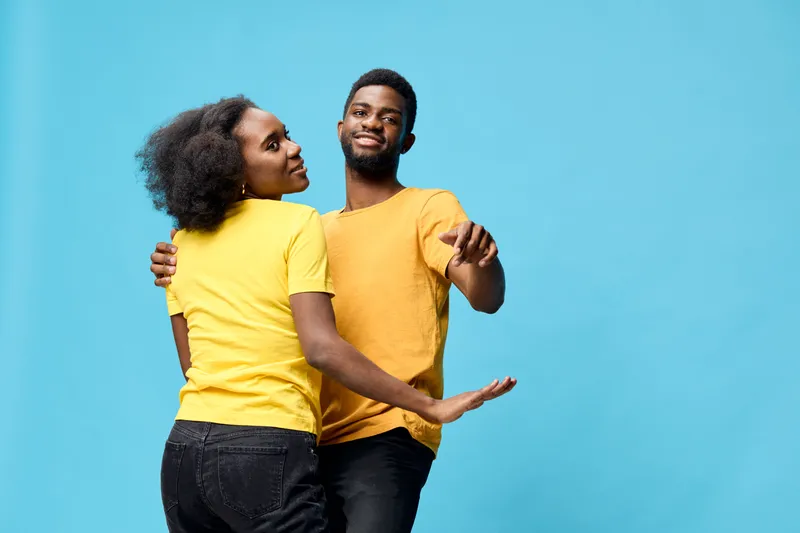 Happy young African couple dancing together against a bright blue background, wearing matching yellow shirts, showcasing joyful emotions and vibrant energy