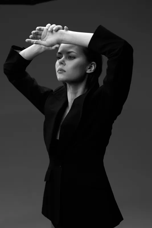 Black and white portrait of a serious woman in a dark blazer posing with arms raised against a dark background.