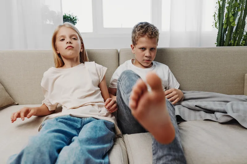 Two children sitting on a beige sofa in a bright living room look serious and relaxed. The boy stretches his foot towards the camera while the girl leans back in casual clothes.