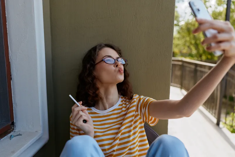 Casual woman takes a selfie on a balcony, wearing a striped shirt and glasses, holding a phone, relaxed and modern, showcasing everyday home lifestyle and outdoor light.