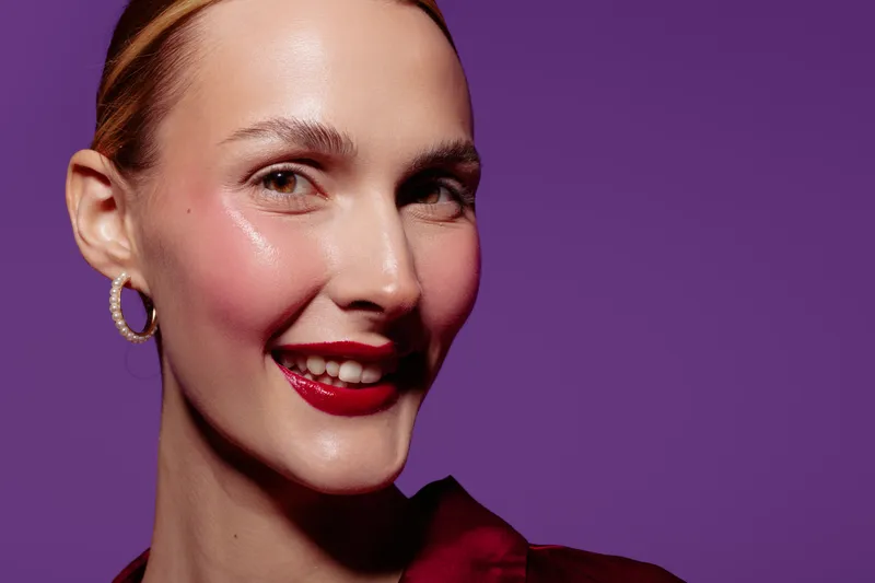 Close-up of smiling woman with red lipstick and hoop earrings on purple background. Concept of beauty, makeup, fashion, and confident female portrait with studio lighting.