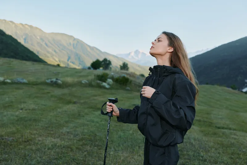 A lone hiker stands in a grassy alpine meadow, gazing upward as she holds a trekking pole. Rugged mountains rise behind her, inviting adventure and exploration in the outdoors.