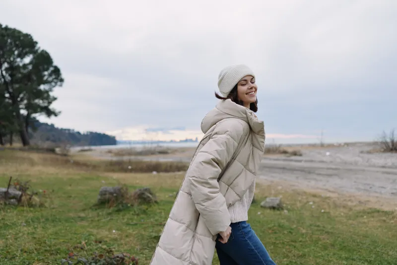 Woman in a beige puffer coat and beanie walks outdoors along a windy coast, showcasing casual winter fashion, comfort and a relaxed, scenic mood.