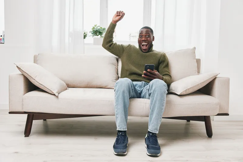 Happy young African American man celebrating success on his smartphone while sitting on a modern couch in a bright room