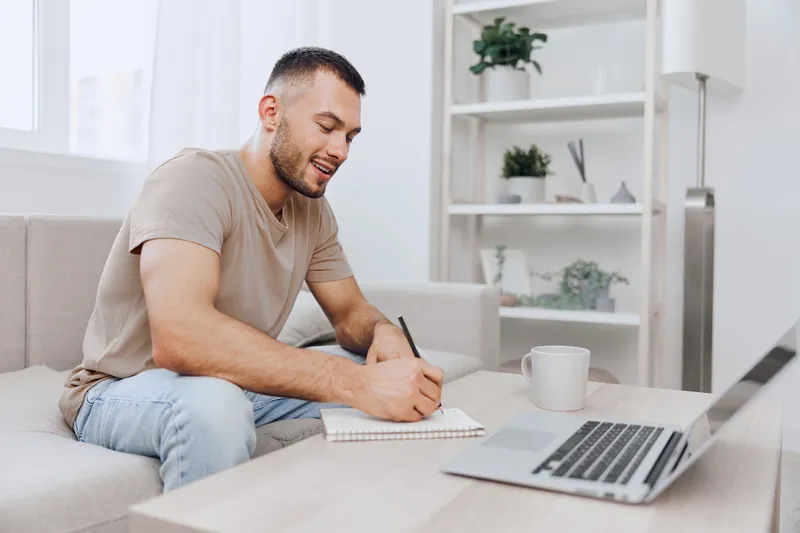 Happy man working from home, engaged in creative writing at a cozy desk. The modern interior and natural light enhance the relaxed atmosphere of productivity.