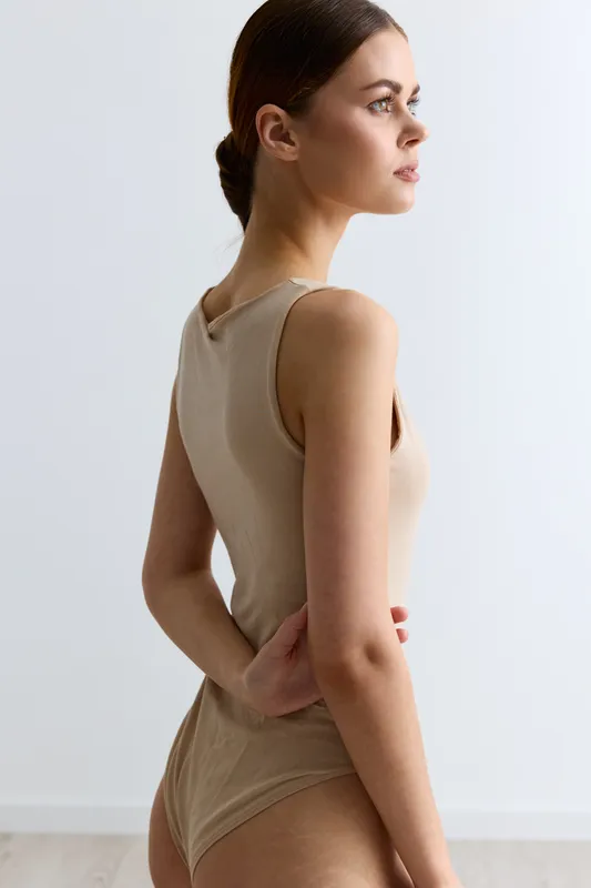 young woman in beige bodysuit posing sideways with focused expression, natural light, minimalist background, soft shadows enhance delicate skin tone