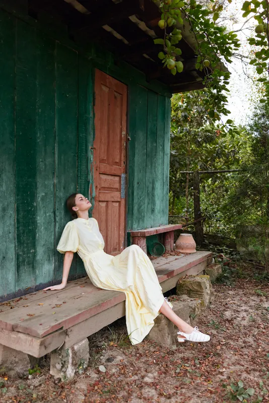 A relaxed woman in a pale yellow dress lounges on a wooden bench beside a green shed, enjoying warm sunlight and a peaceful countryside atmosphere.